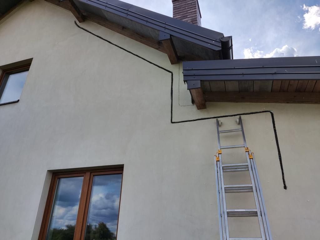 Elewacja domu z poprowadzonymi przewodami elektrycznymi w czarnych rurkach, drabina oparta o ścianę, widoczne okna z drewnianą ramą i fragment dachu z rynną.