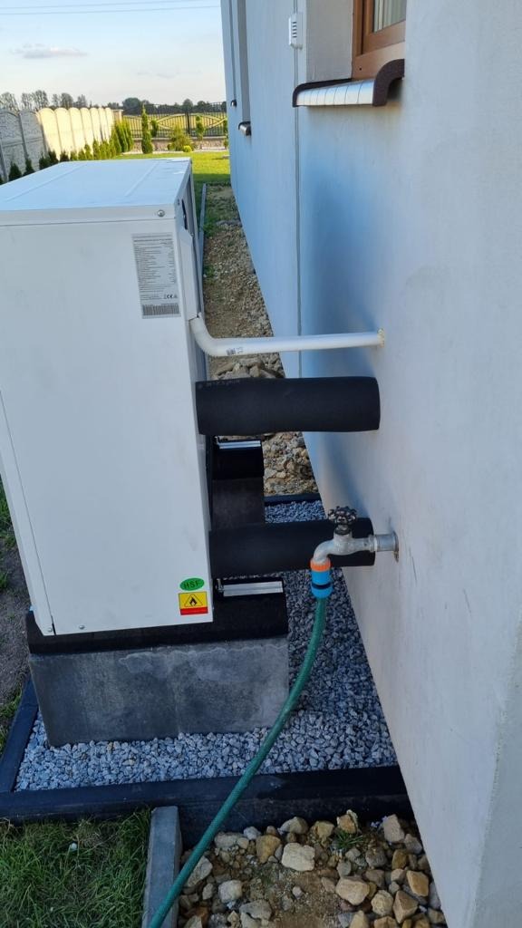 Zewnętrzna jednostka pompy ciepła na betonowym fundamencie, z izolowanymi rurami przy ścianie budynku i podłączonym wężem ogrodowym do zaworu.