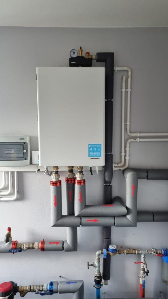 Biała obudowa urządzenia grzewczego z podłączonymi rurami izolowanymi szarą pianką z czerwonymi strzałkami, manometr na górze, instalacja w pomieszczeniu o szarych ścianach.