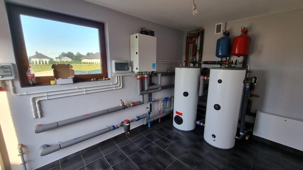 Instalacja w kotłowni: dwa białe zasobniki ciepłej wody z niebieskim i czerwonym zbiornikiem wyrównawczym, szare rury instalacyjne, biały piec wiszący na ścianie. W tle okno z widokiem na domy.