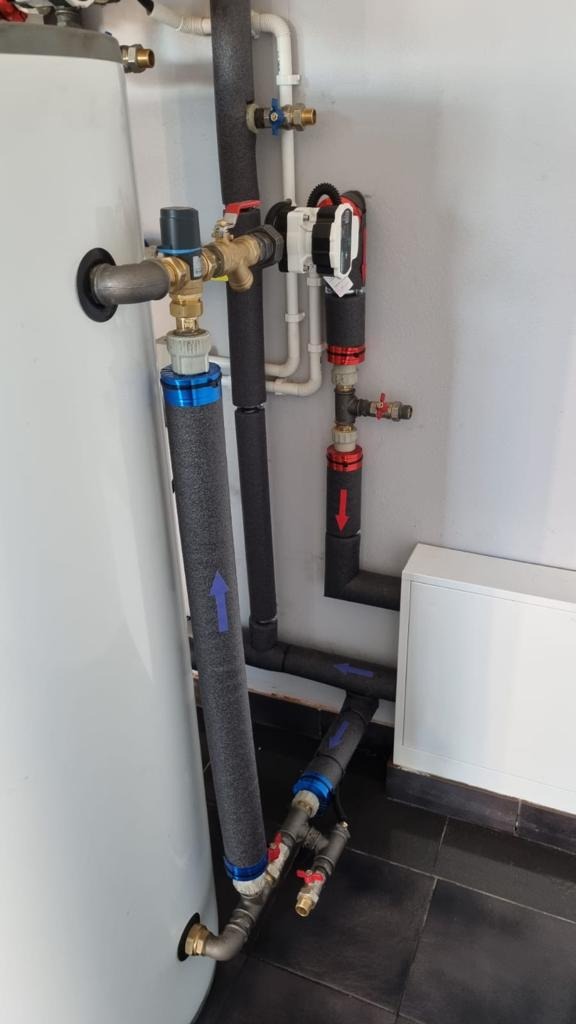 Instalacja grzewcza z rurami w izolacji, zaworami i pompą obiegową w kotłowni z białymi ścianami i ciemną podłogą z płytek. Na izolacji rur widoczne niebieskie strzałki wskazujące kierunek przepływu.