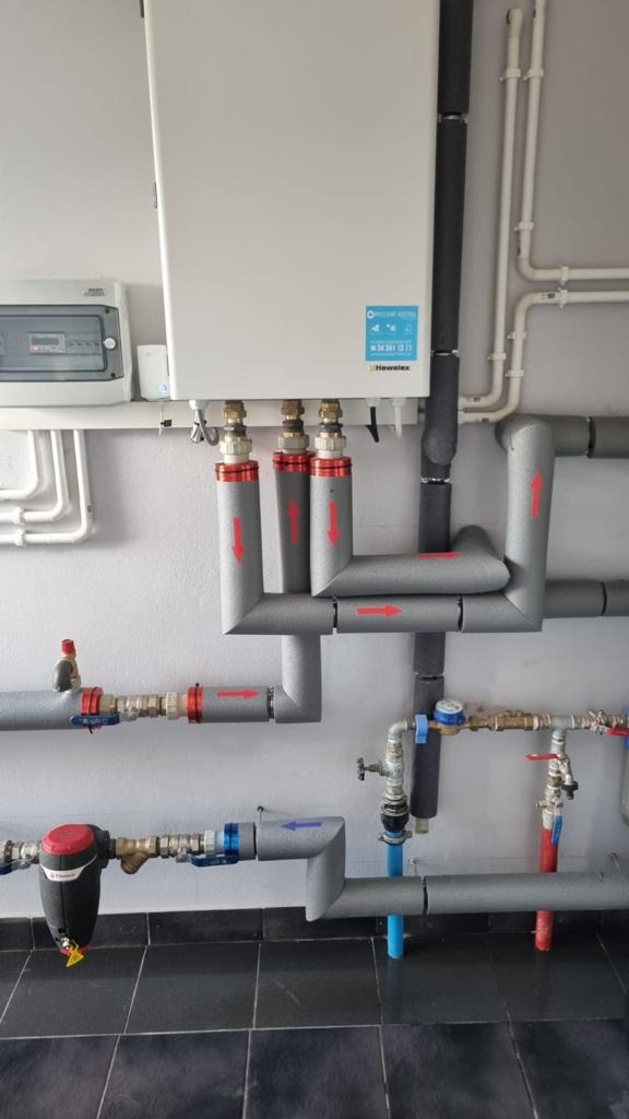 Instalacja grzewcza z białym zasobnikiem ciepłej wody, szare rury z izolacją i czerwonymi strzałkami wskazującymi kierunek przepływu, oraz licznikiem zużycia na czarnej podłodze.