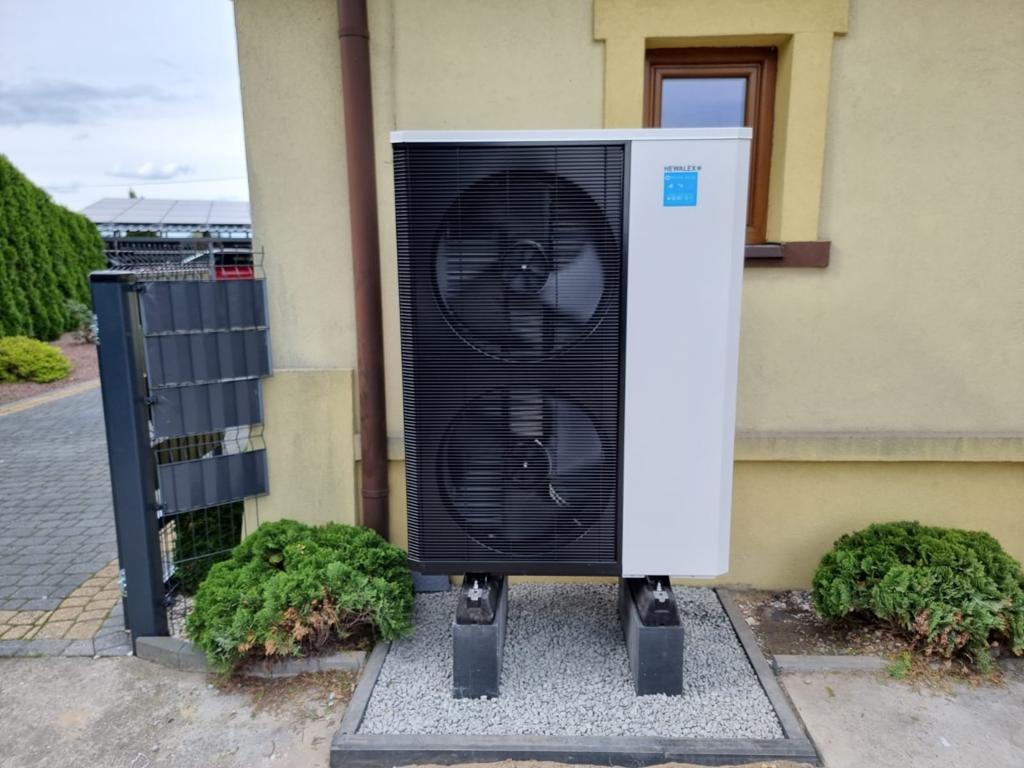 Biała pompa ciepła z czarnym grillem wentylacyjnym, umieszczona na betonowych blokach przed ścianą budynku z brązowym oknem, obok rynny spustowej i niskiego żywopłotu. W tle widoczne panele...