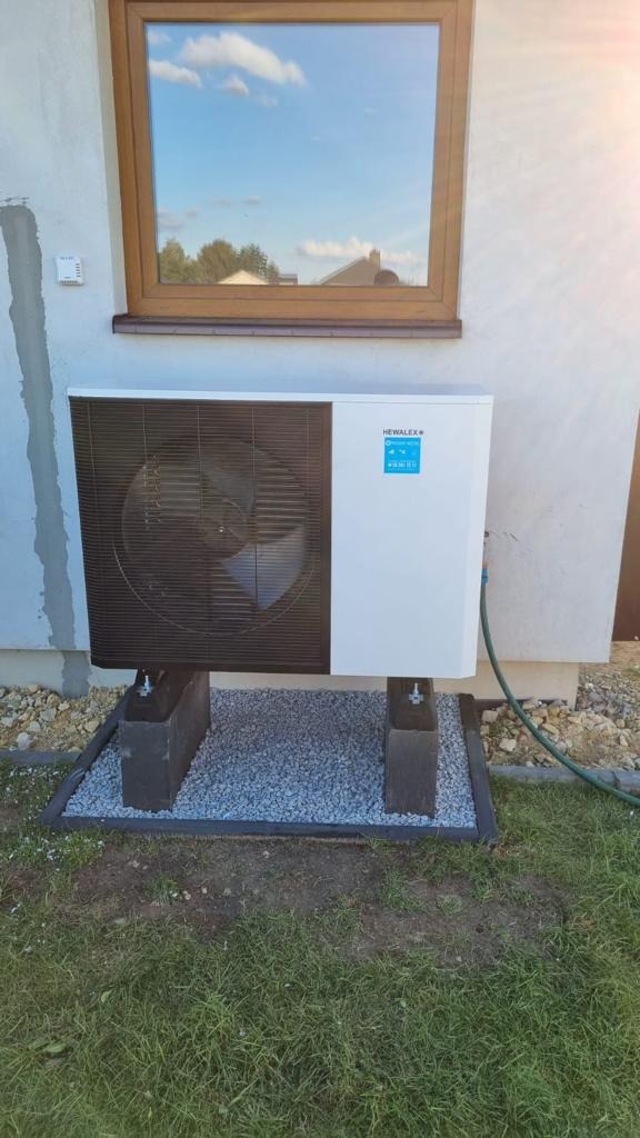 Zewnętrzna jednostka pompy ciepła Hewalex na betonowych wspornikach, z widocznym oknem odbijającym błękitne niebo i chmury.