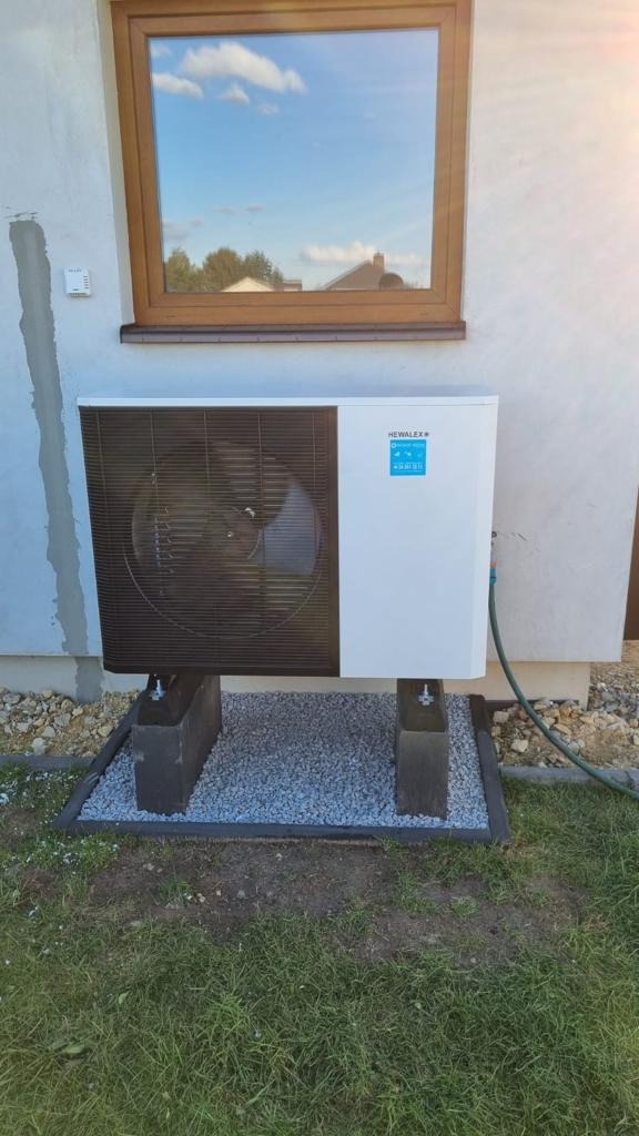 Zewnętrzna jednostka pompy ciepła Hewalex zainstalowana na betonowych podstawach z podsypką z drobnego żwiru, widoczna elewacja budynku z oknem odbijającym niebo.