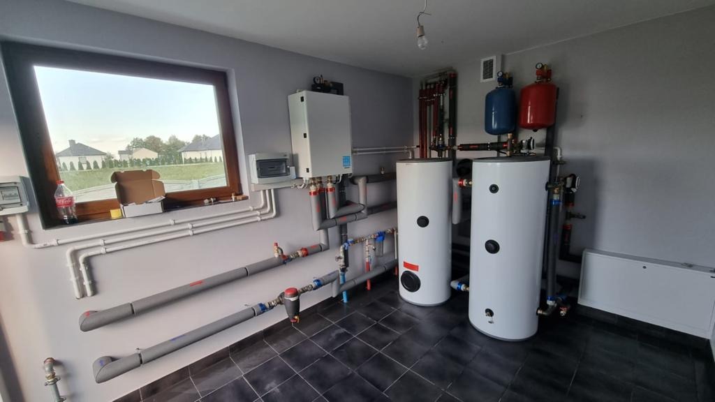 Wnętrze kotłowni z dwoma białymi zbiornikami akumulacyjnymi, wiszącym kotłem gazowym, rozbudowaną instalacją hydrauliczną z rurami w izolacji, okno z widokiem na domy jednorodzinne.