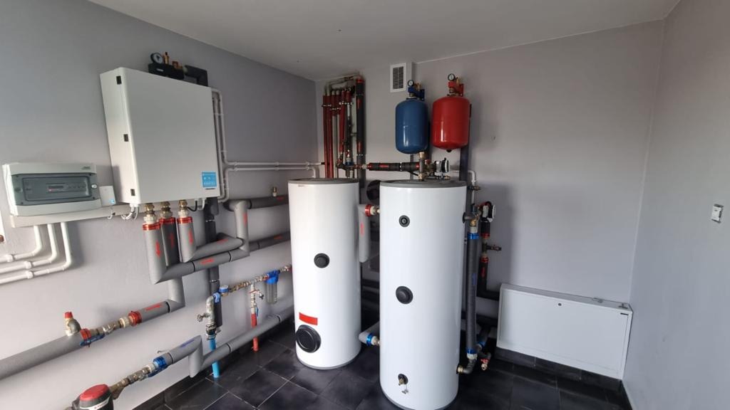 Zestaw dwóch białych zbiorników akumulacyjnych ciepła z niebieskim i czerwonym zbiornikiem wyrównawczym, połączonych rurami w kotłowni z szarymi ścianami i czarną podłogą.