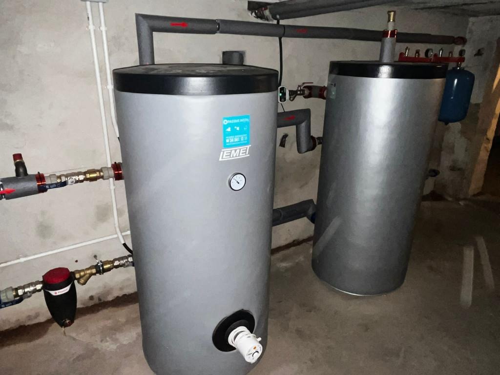 Dwa szare zbiorniki akumulacyjne wody użytkowej z czarnymi pokrywami, podłączone do rur w piwnicy, z widocznymi zaworami i manometrem na jednym z nich.