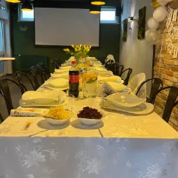 Uroczysty stół komunijny z białym obrusem w kwiaty, zastawą, sałatkami i napojami, dekorowany balonami i girlandą na tle ceglanej ściany.