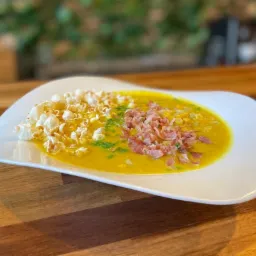 Kremowa zupa kukurydziana na białym talerzu, udekorowana popcornem, smażonym boczkiem i świeżym szczypiorkiem, na drewnianym stole.
