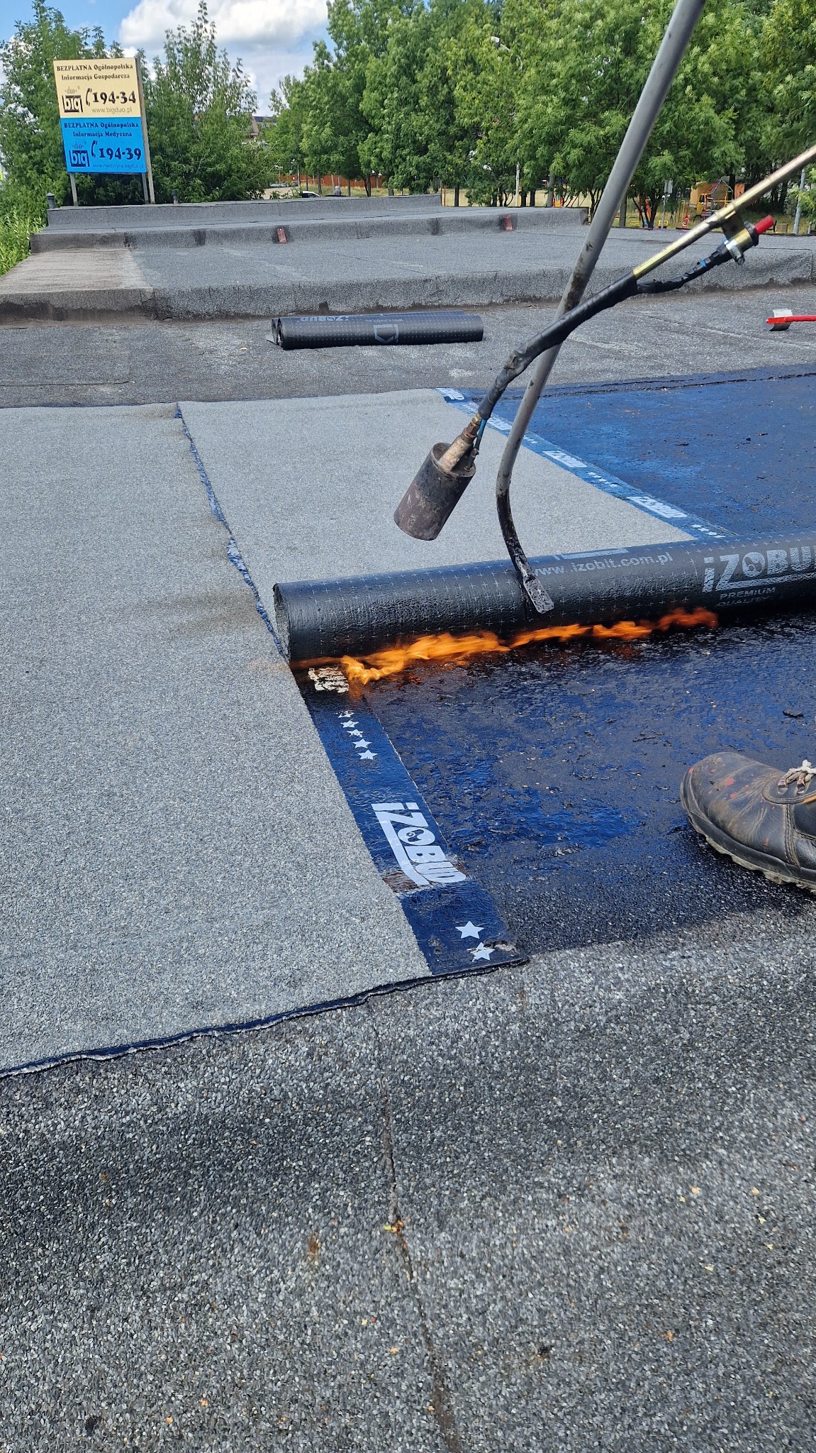 Krycie dachu papą termozgrzewalną z użyciem palnika. Widoczny fragment dachu, rolka papy Izobud i płomień palnika. W tle budynki i zieleń.