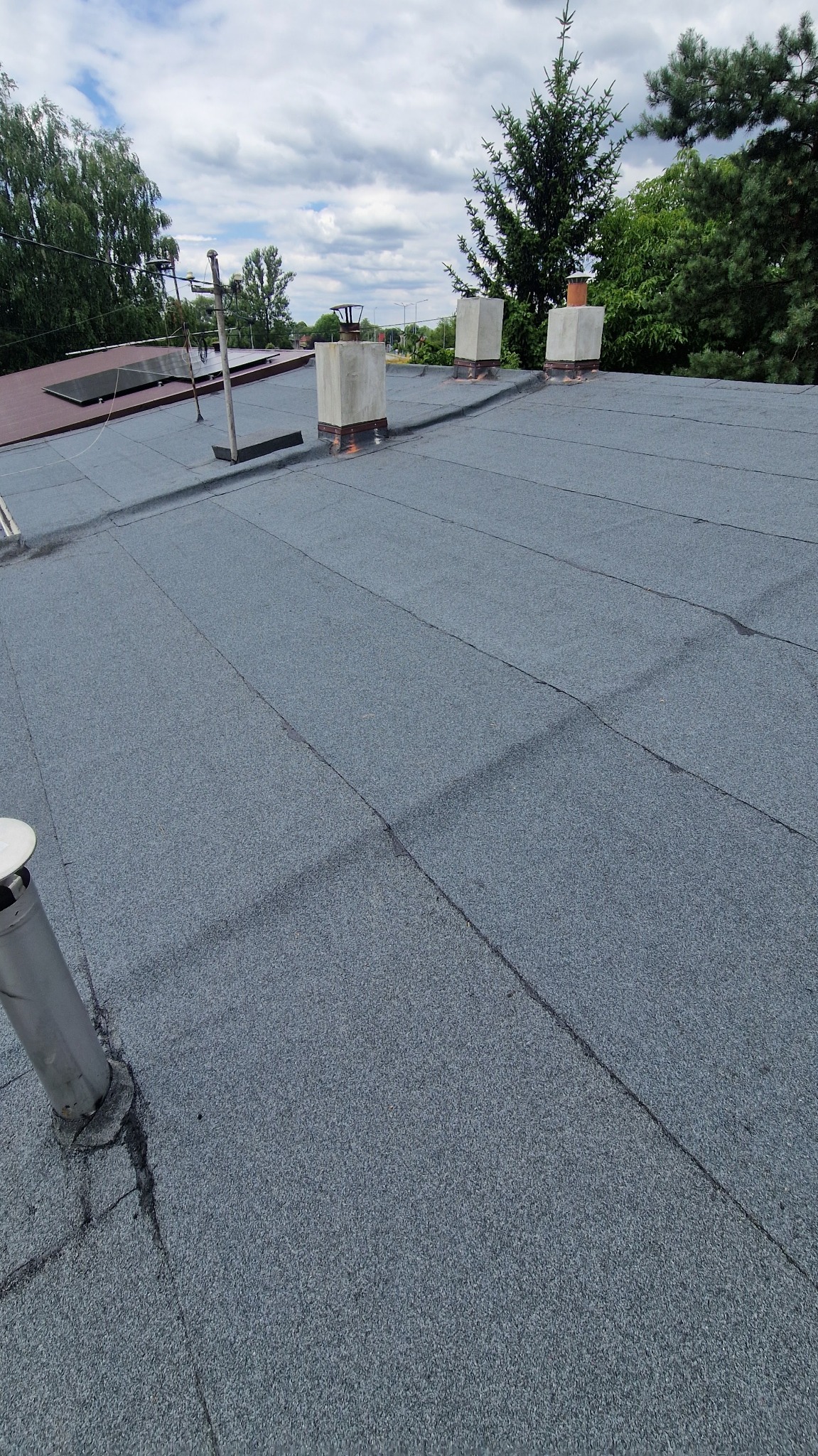 Szary, płaski dach pokryty papą z trzema kominami i elementami instalacji odgromowej. Widok z góry, w tle zieleń drzew i błękitne niebo.