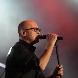 Mężczyzna w okularach, śpiewający do mikrofonu na scenie z czerwono-białym oświetleniem.