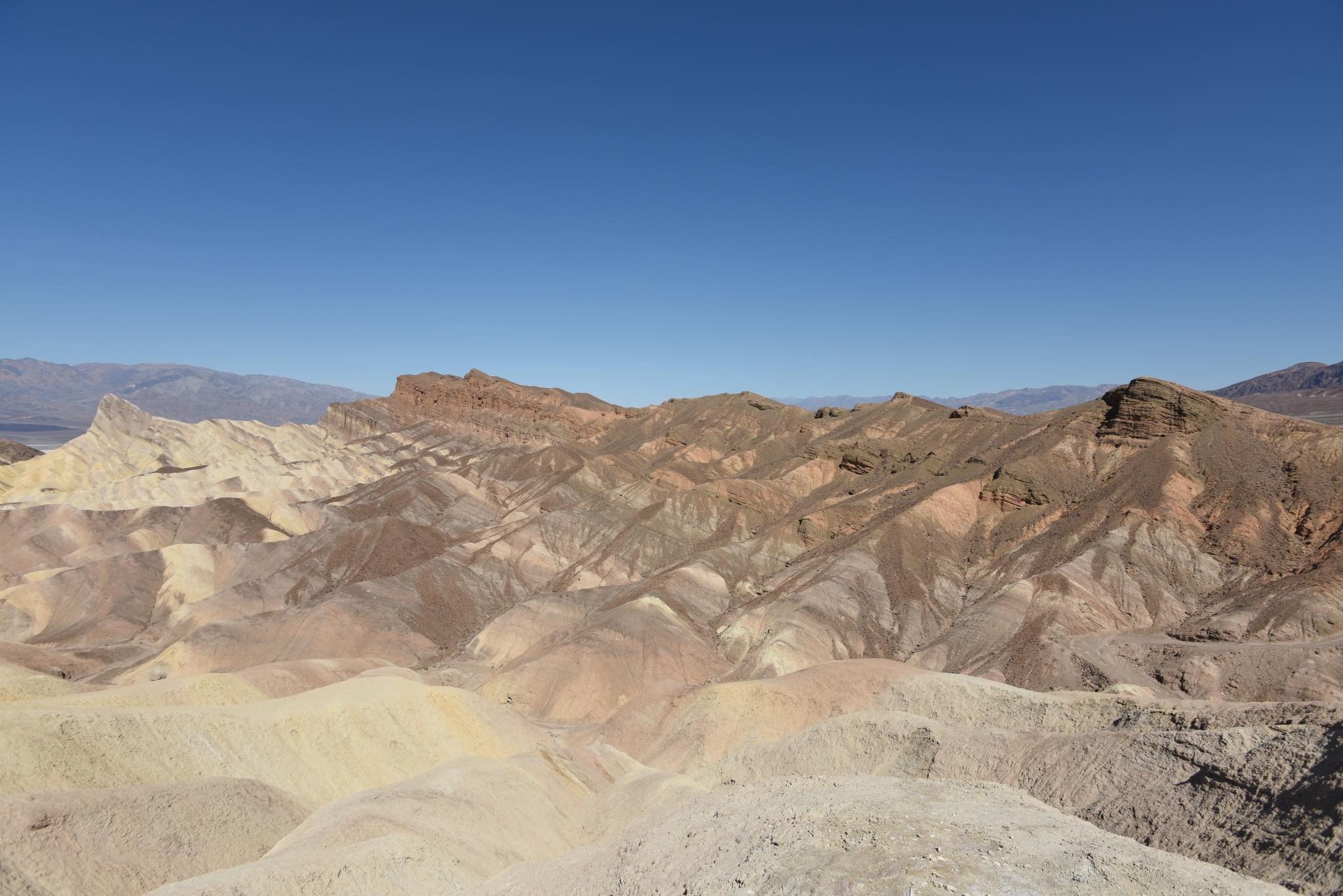 Panoramiczny widok na pustynny krajobraz z formacjami skalnymi w odcieniach beżu, brązu i szarości pod błękitnym niebem, z górami widocznymi w oddali.