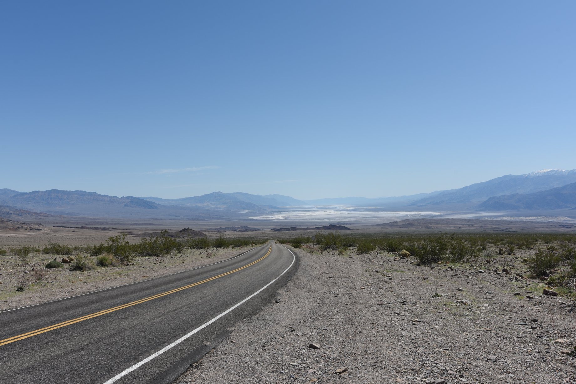 Asfaltowa droga z podwójną żółtą linią i pojedynczą białą linią prowadząca przez pustynny krajobraz z górami w tle pod błękitnym niebem.