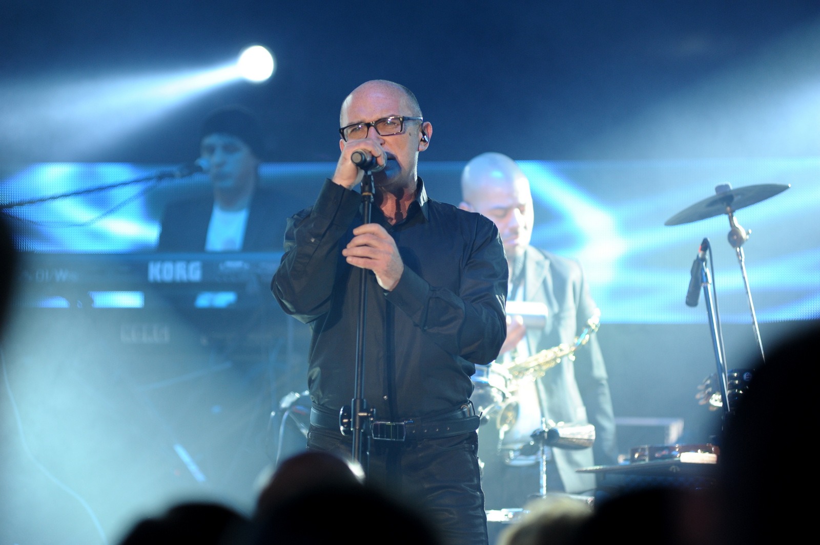 Wokalista w czarnej koszuli i okularach śpiewa do mikrofonu na scenie, w tle keyboard Korg, saksofonista i perkusja, oświetlenie sceniczne.