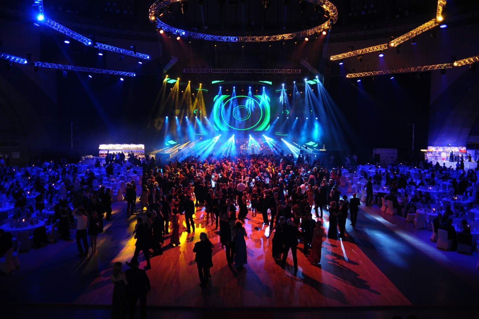 Szeroki kadr na salę balową wypełnioną gośćmi, z podświetloną sceną i dekoracjami świetlnymi w kolorach niebieskim i żółtym.