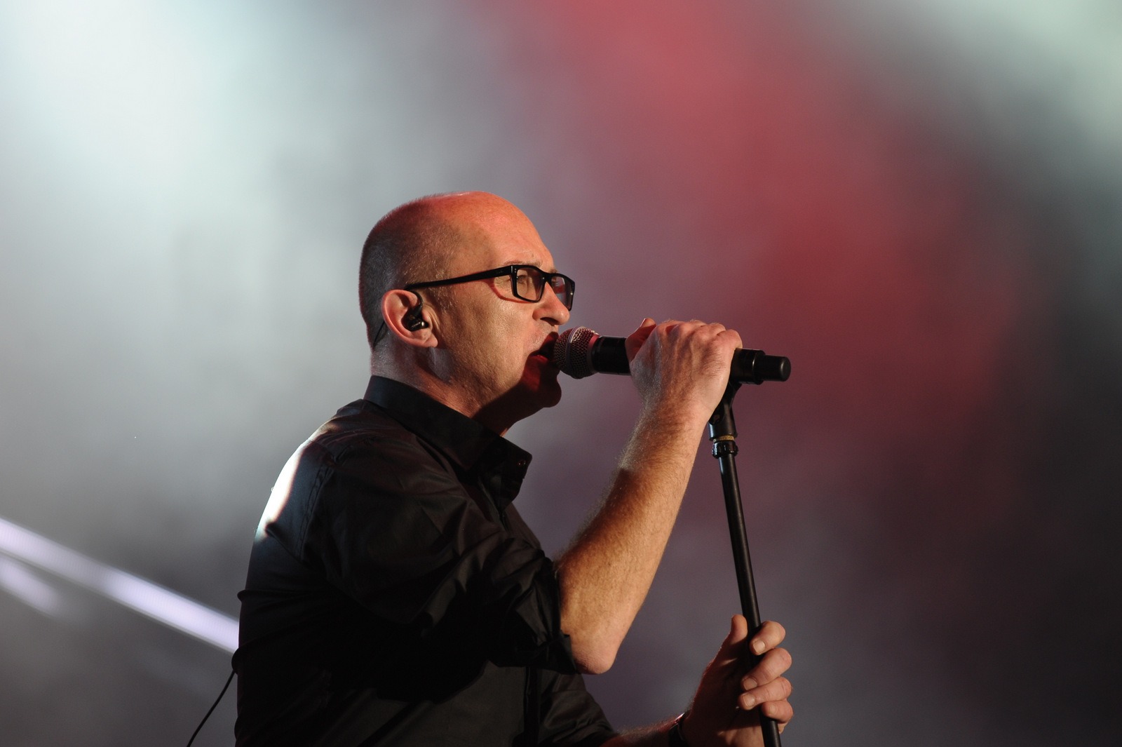 Mężczyzna w okularach, śpiewający do mikrofonu na scenie z czerwono-białym oświetleniem.