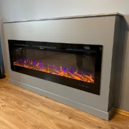 Szary kominek elektryczny z efektem płomieni w zabudowie, widok z boku na drewnianą podłogę i fragment ściany z obrazem.