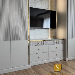 Szara szafa wnękowa z frezowanymi frontami i komodą pod telewizor, wykończona złotymi detalami na tle marmurowej ściany.