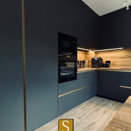 SKOWROŃSKI Kuchnie&Meble na wymiar - Nowoczesna, grafitowa kuchnia z drewnianym akcentem nad blatem i złotymi detalami. Widoczne sprzęty AGD i dekoracje. Podłoga drewniana, ułożona w jodełkę.
