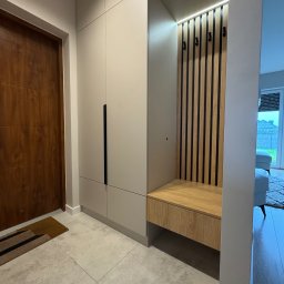 SKOWROŃSKI Kuchnie&Meble na wymiar - Beżowa szafa w przedpokoju z siedziskiem i wieszakami na drewnianym panelu. Nowoczesny design, minimalistyczne uchwyty, jasne płytki podłogowe.
