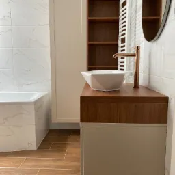 Nowoczesna łazienka z białą umywalką nablatową o geometrycznym kształcie, złotą armaturą, szafką pod umywalką w odcieniach beżu i drewna, oraz białymi płytkami imitującymi marmur na ścianach.