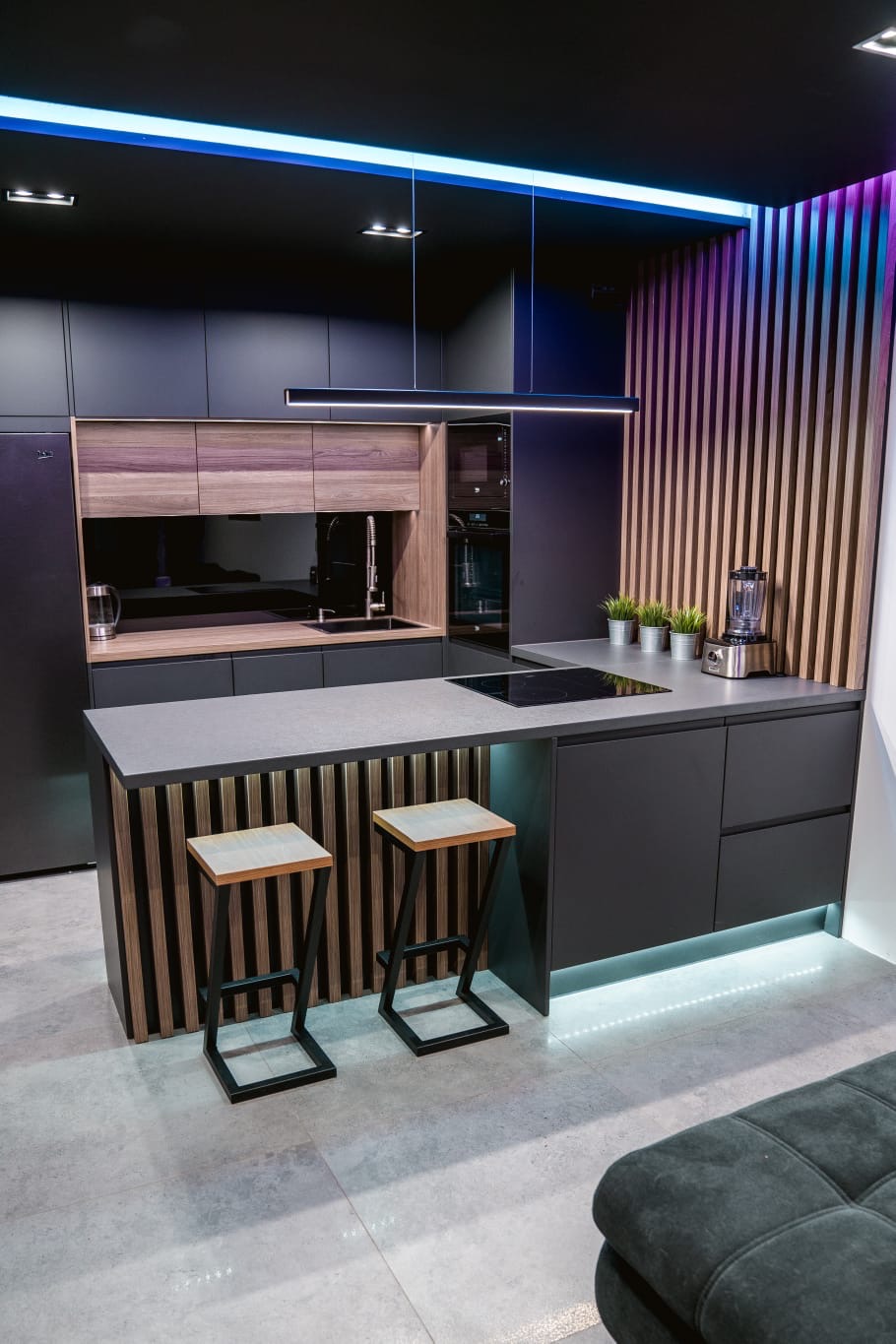 Nowoczesna kuchnia z czarnymi szafkami, drewnianymi akcentami i podświetleniem LED, widoczne dwa stołki barowe z drewnianymi siedziskami i metalowymi nogami.