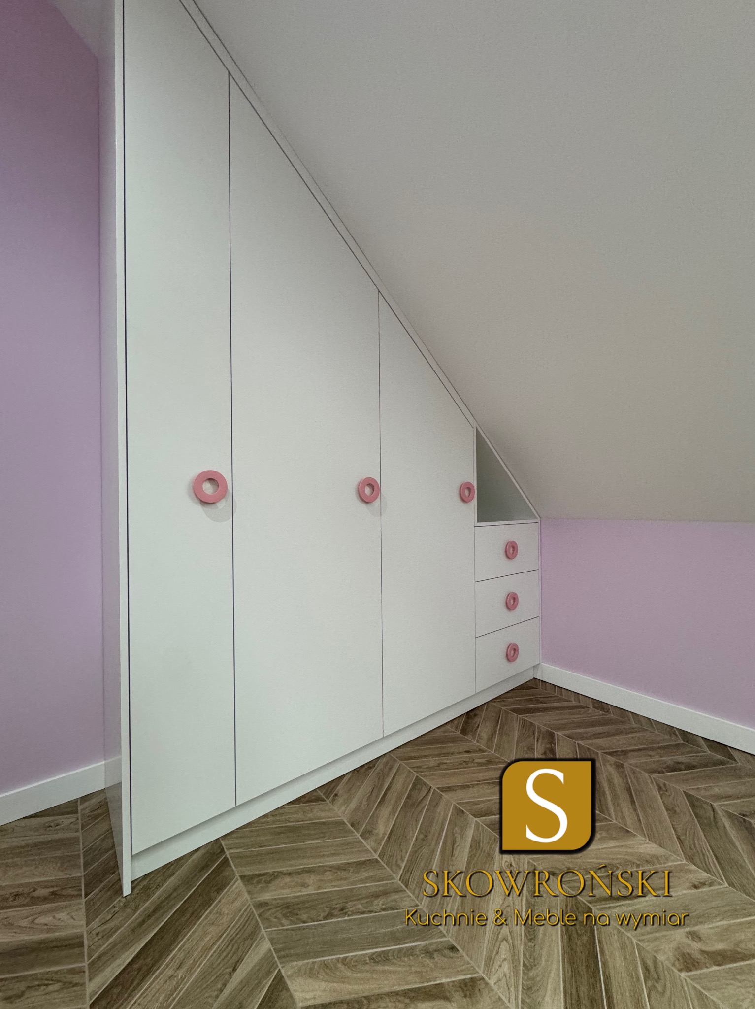 Biała szafa wnękowa na poddaszu ze skośnym sufitem, z trzema drzwiami i trzema szufladami, różowe okrągłe uchwyty, parkiet w jodełkę na podłodze.