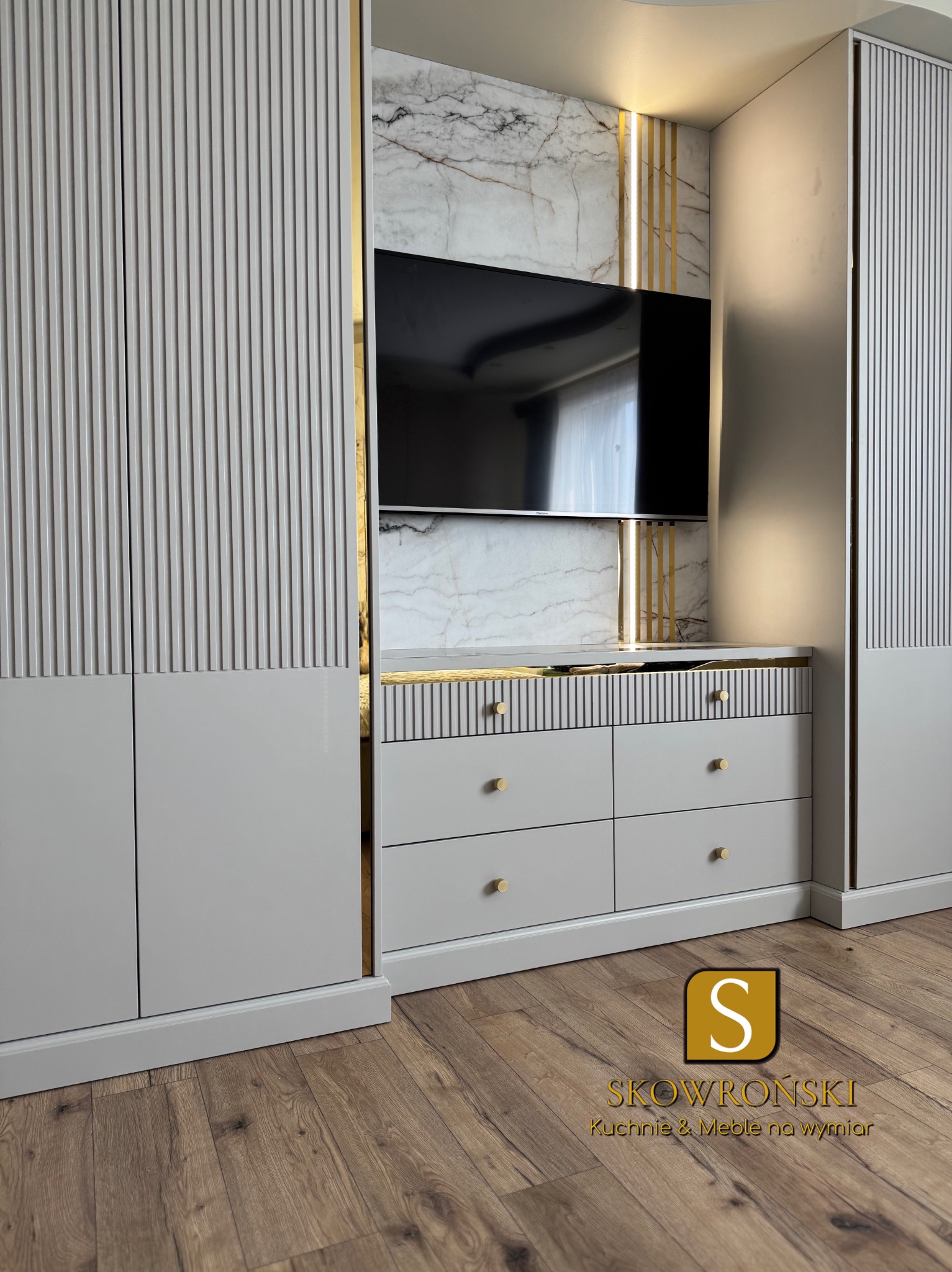 Szara szafa wnękowa z frezowanymi frontami i komodą pod telewizor, wykończona złotymi detalami na tle marmurowej ściany.