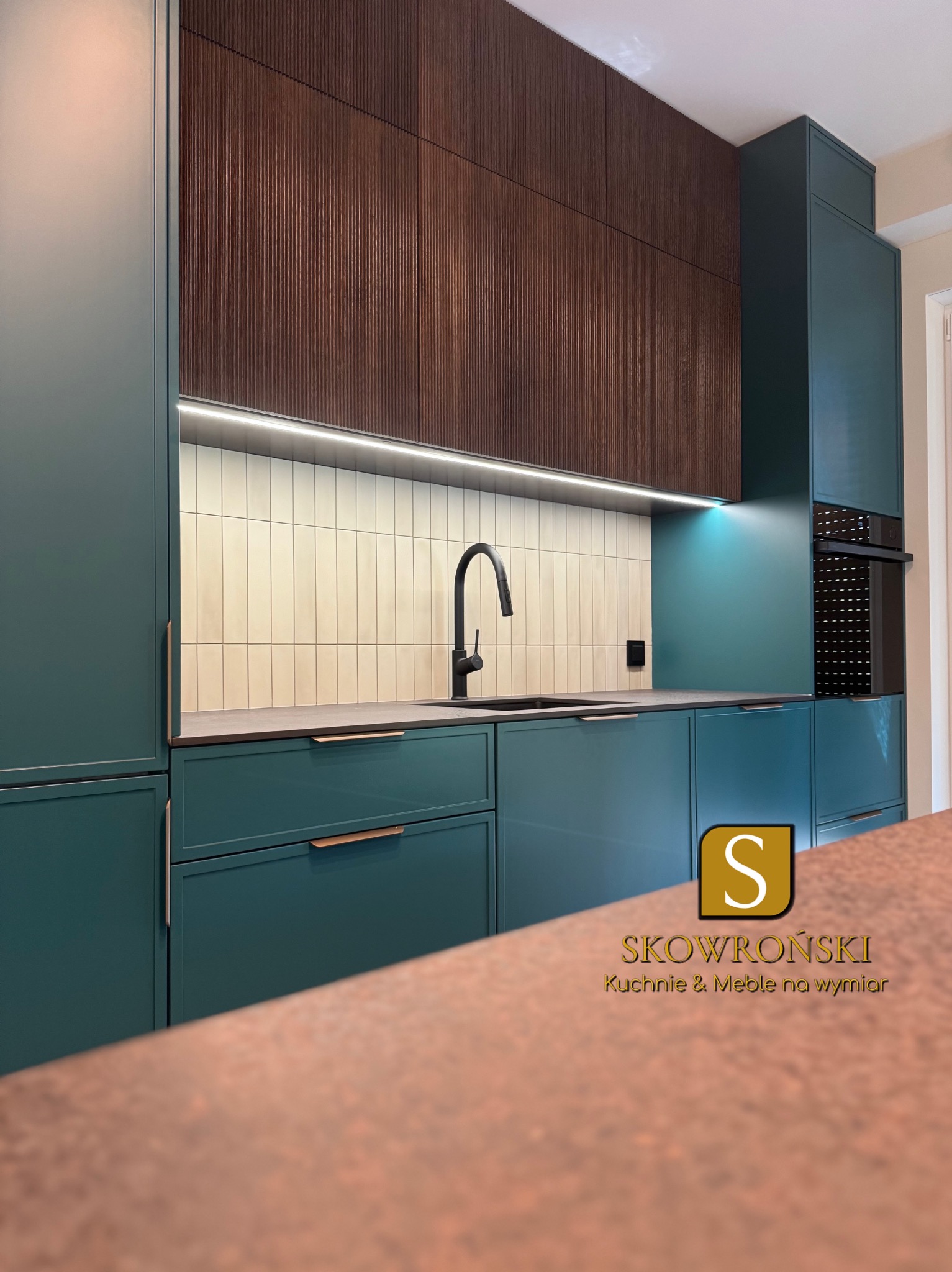 Nowoczesna kuchnia z zielonymi szafkami i drewnianymi frontami górnymi. Uwagę przykuwa czarna bateria i subtelne oświetlenie LED pod szafkami.