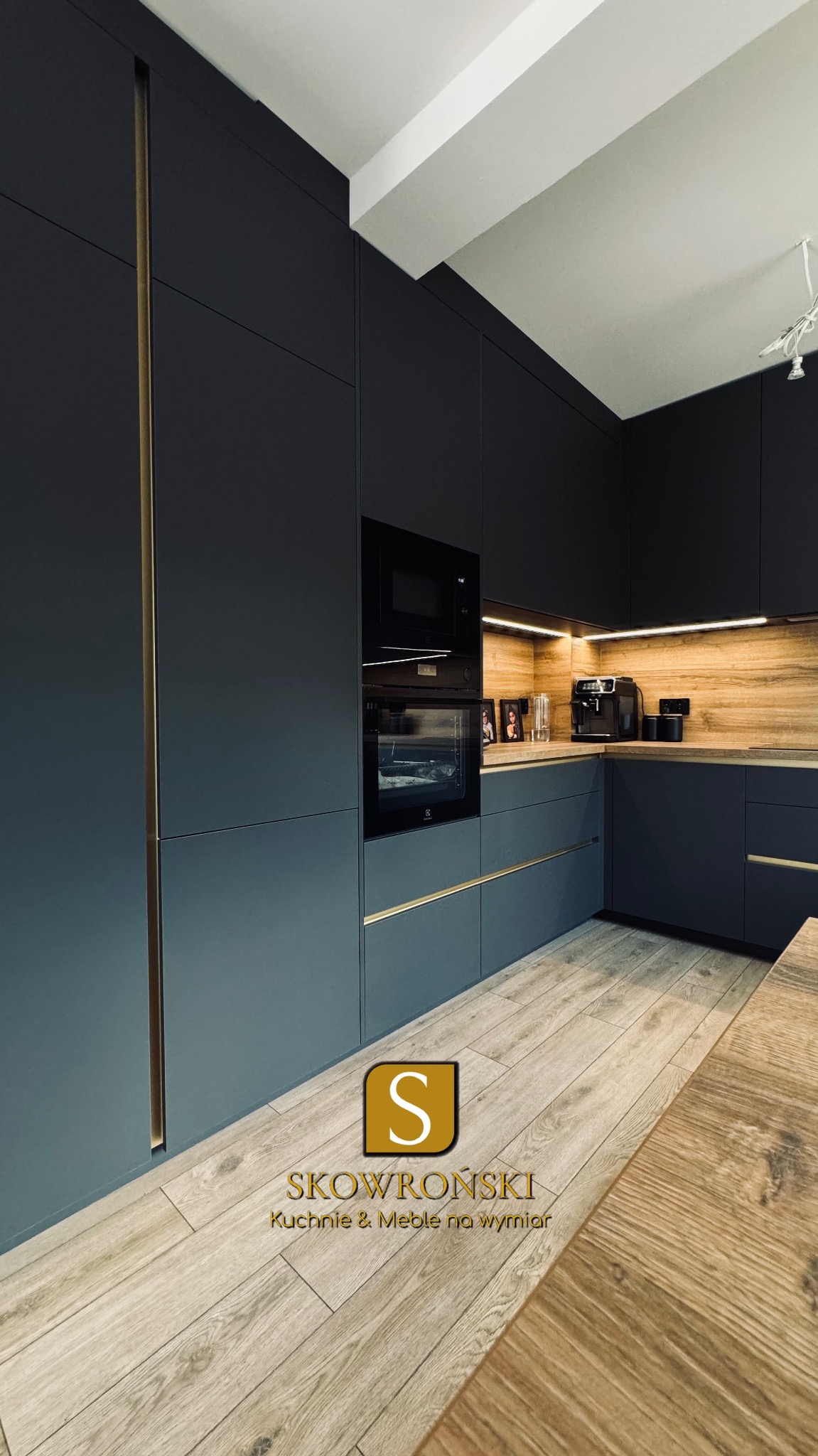Nowoczesna, grafitowa kuchnia z drewnianym akcentem nad blatem i złotymi detalami. Widoczne sprzęty AGD i dekoracje. Podłoga drewniana, ułożona w jodełkę.