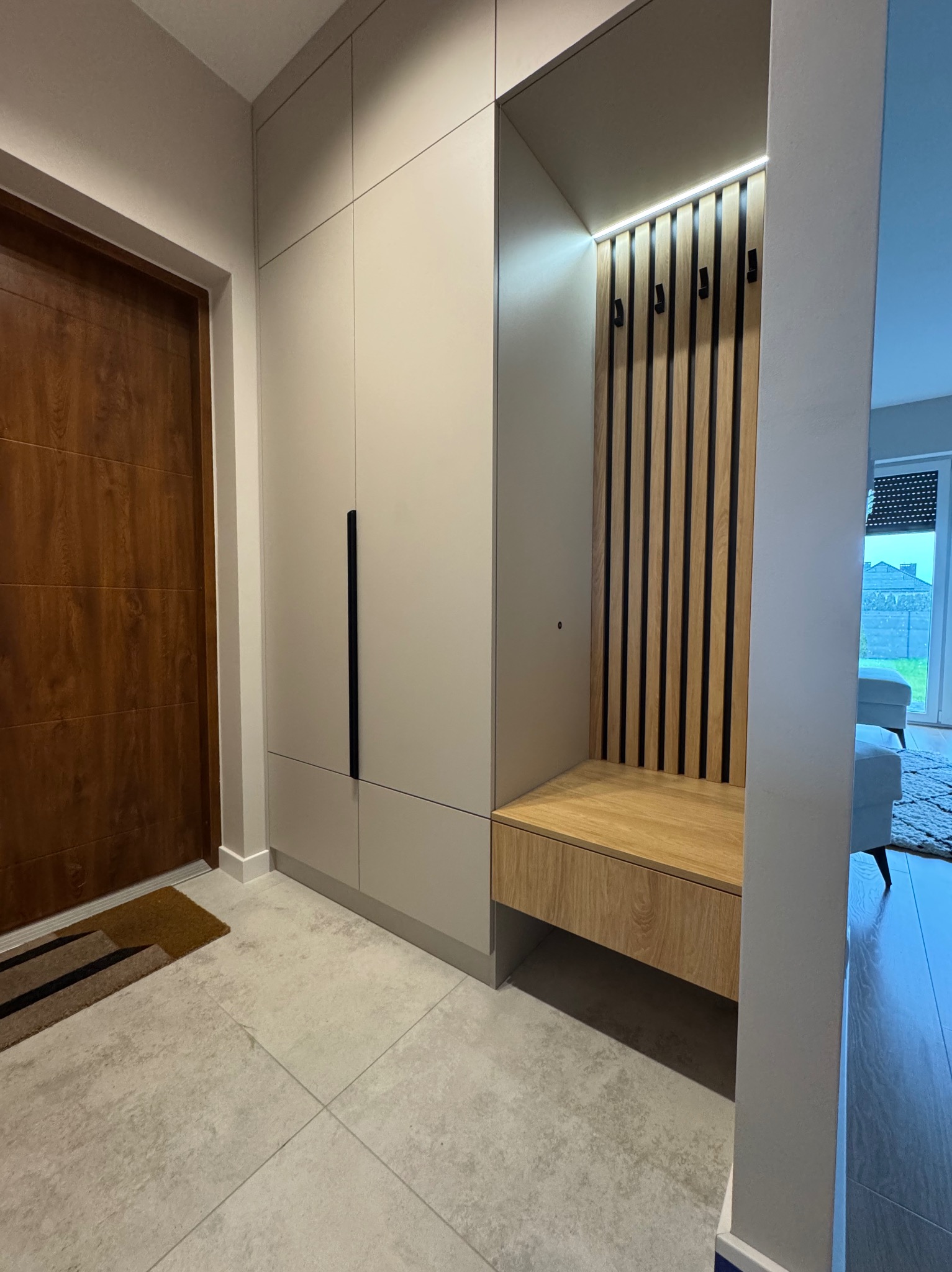 Beżowa szafa w przedpokoju z siedziskiem i wieszakami na drewnianym panelu. Nowoczesny design, minimalistyczne uchwyty, jasne płytki podłogowe.