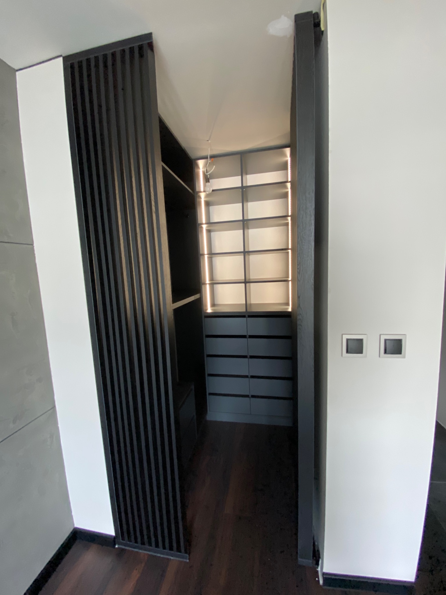 Nowoczesna szafa wnękowa z podświetlanymi półkami i ciemnymi szufladami, widoczna przez dekoracyjne, pionowe lamele w Poznaniu.