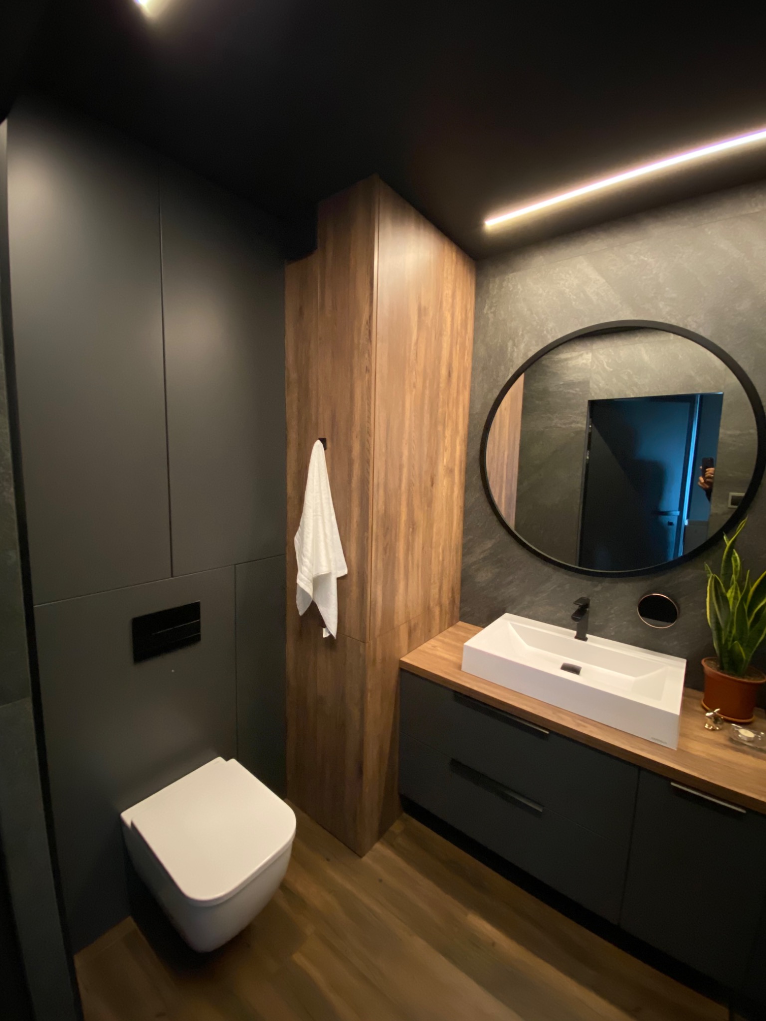 Nowoczesna łazienka w odcieniach szarości i drewna z podwieszaną toaletą, umywalką nablatową i okrągłym lustrem z czarną ramą.