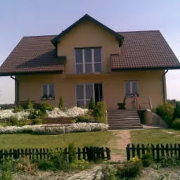 Dwukondygnacyjny dom z beżową elewacją i brązowym dachem, otoczony ogrodem z białymi kwiatami i drewnianym płotem.