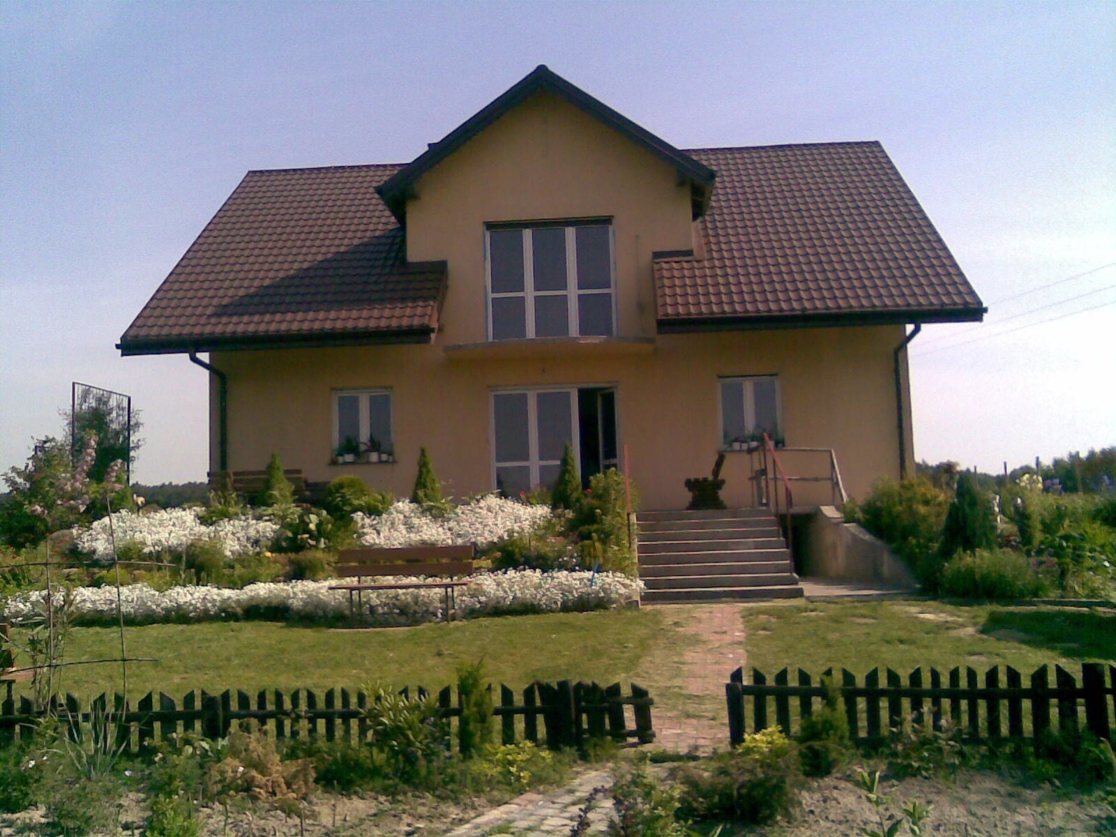 Dwukondygnacyjny dom z beżową elewacją i brązowym dachem, otoczony ogrodem z białymi kwiatami i drewnianym płotem.