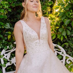 Katarzyna Sałata Fotografia - Panna młoda w sukni ślubnej z głębokim dekoltem, siedząca na białej ławce w ogrodzie, z zamkniętymi oczami i uśmiechem, w promieniach słońca.