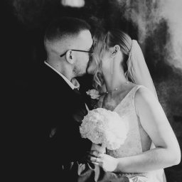 Katarzyna Sałata Fotografia - Czarno-białe zbliżenie całującej się pary młodej. Panna młoda trzyma bukiet białych kwiatów. W tle delikatne rozmycie, nadające scenie romantyczny charakter.