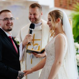 Katarzyna Sałata Fotografia - Para młoda trzymająca się za ręce podczas ceremonii ślubnej w kościele, z uśmiechniętym księdzem w tle, z widocznym tatuażem na dłoni pana młodego.