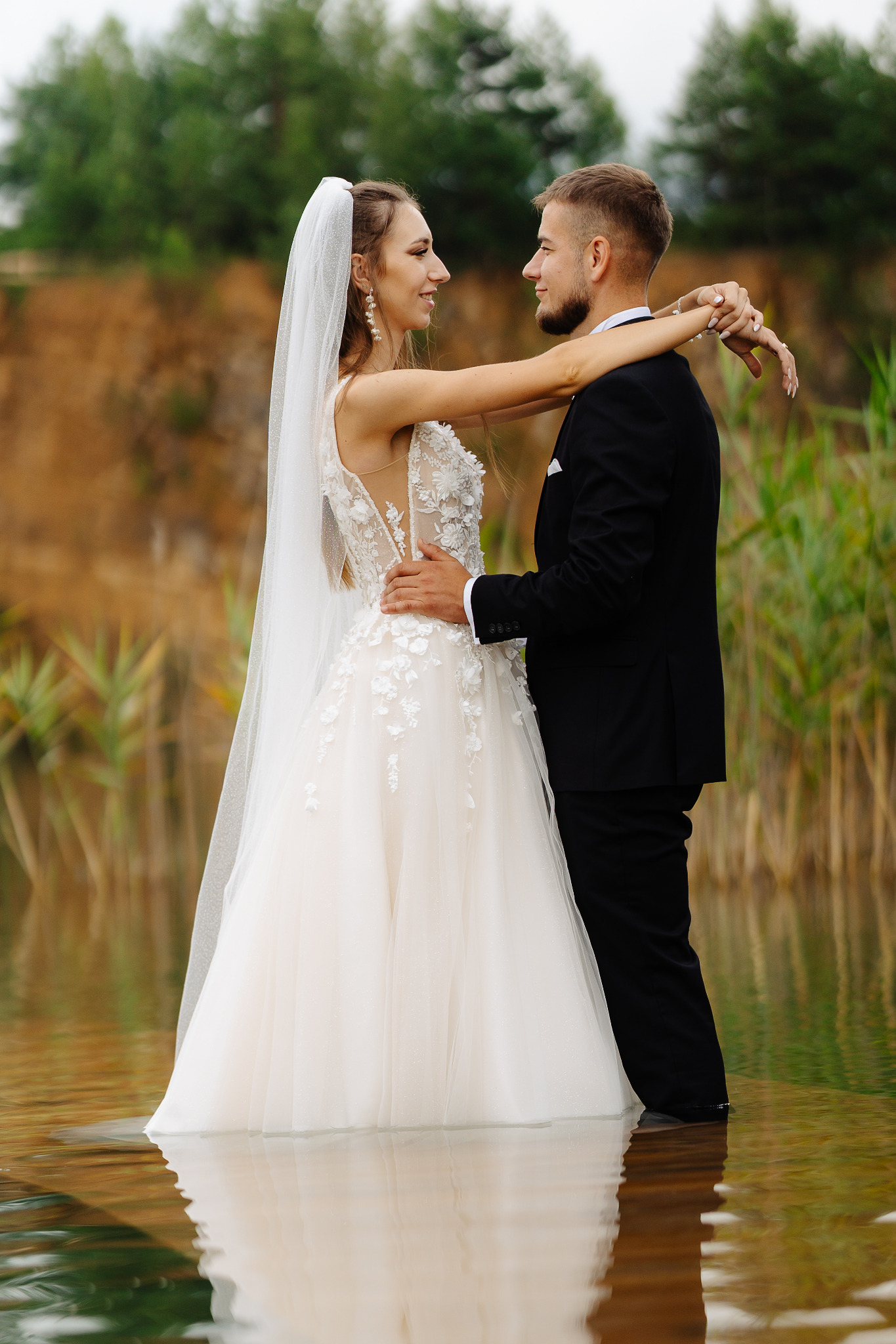 Para młoda w wodzie, on w czarnym garniturze, ona w białej sukni z welonem. Uśmiechnięci, patrzą na siebie, w tle zielona roślinność i piaszczysty brzeg.