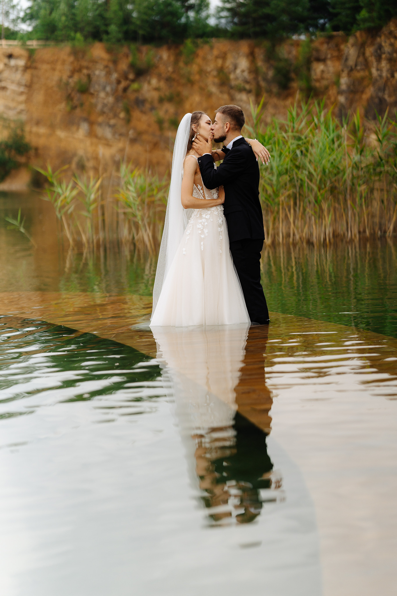 Para młoda w objęciach, pocałunek na tle malowniczego, wodnego krajobrazu z trzcinami i skalistym brzegiem, odbicie w wodzie, suknia z koronką.