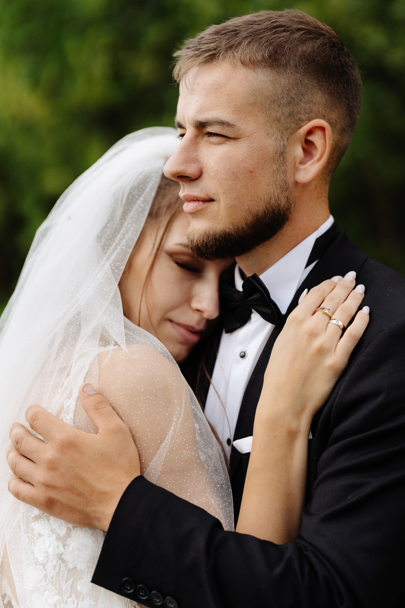Ujęcie obejmującej się pary młodej w plenerze: panna młoda w welonie i sukni ślubnej, pan młody w smokingu z muchą. Widoczne obrączki.