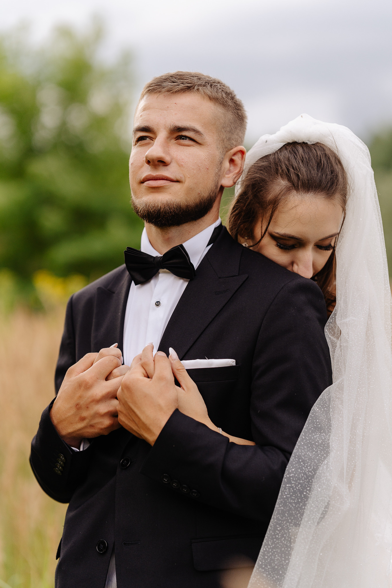 Para młoda w objęciach na tle zieleni. Pan młody w czarnym smokingu z muchą, panna młoda w białej sukni i welonie z delikatnym połyskiem.