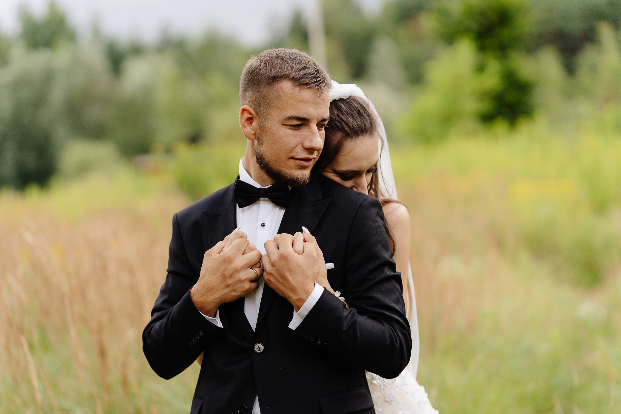 Para młoda w objęciach na tle zieleni. Pan młody w czarnym smokingu z muszką, panna młoda w białej sukni z welonem. Romantyczna sceneria ślubna.
