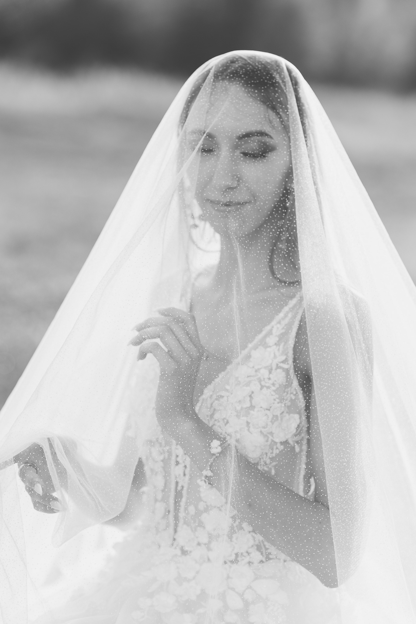 Portret panny młodej w czarno-białej tonacji, z delikatną woalką z kropeczkami. Suknia z koronkowymi aplikacjami. Subtelny uśmiech i zamknięte oczy.