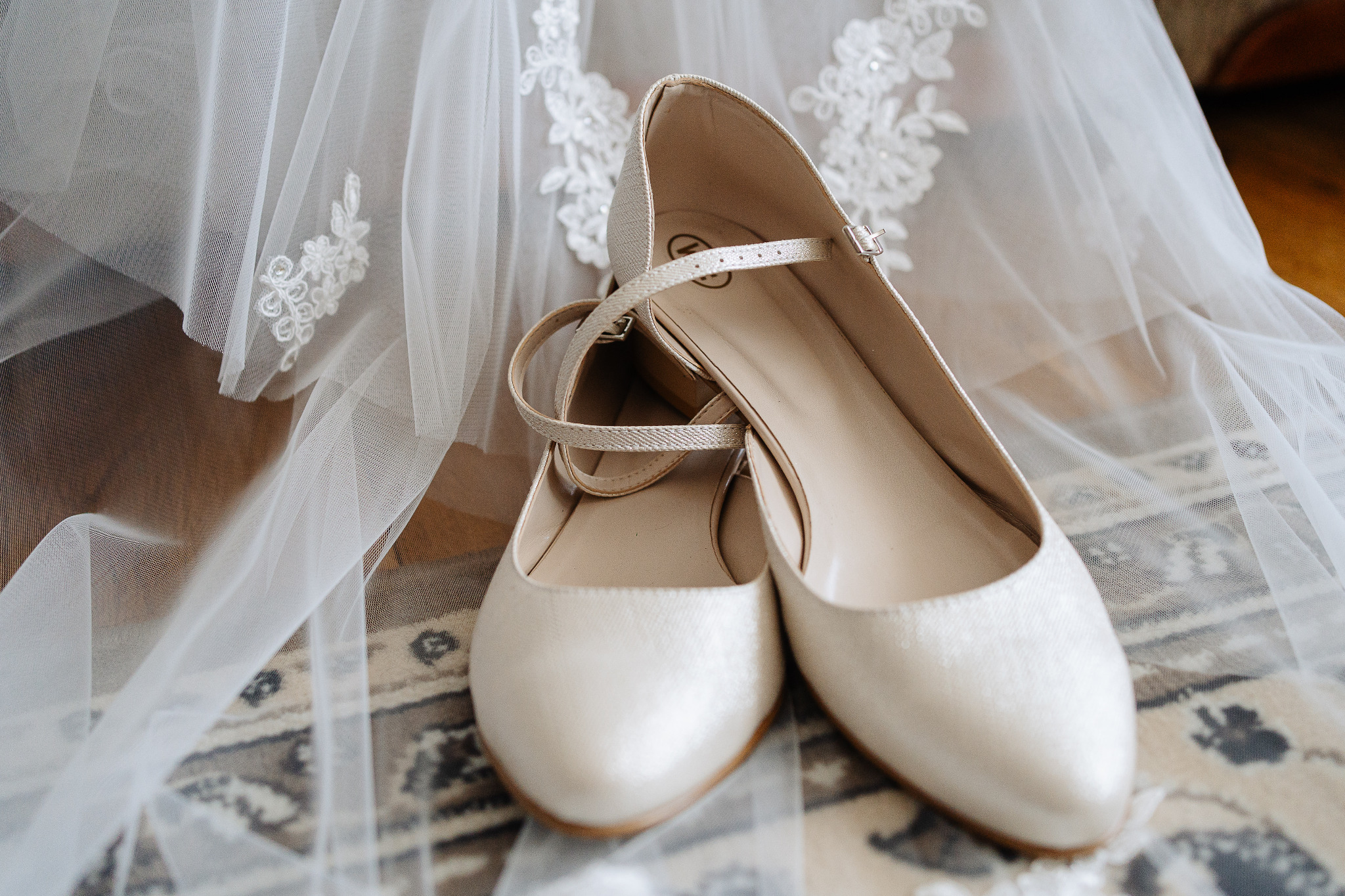 Eleganckie, kremowe baleriny ślubne z paskami, leżące na wzorzystym dywanie obok fragmentu welonu z haftowanymi aplikacjami. Delikatne światło podkreśla detale.