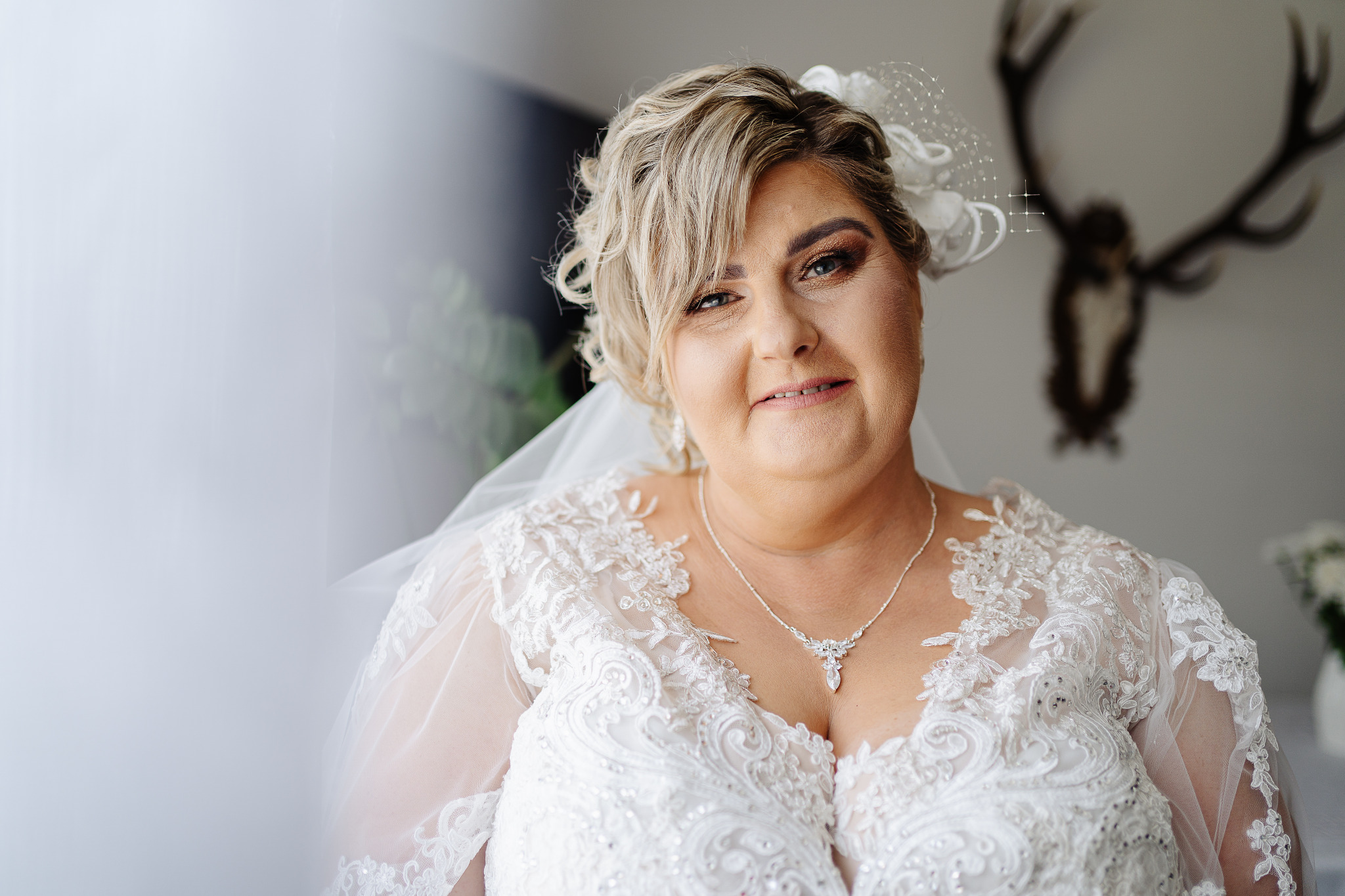 Portret uśmiechniętej panny młodej w koronkowej sukni, z delikatnym makijażem i welonem, na tle ściany z porożem jelenia. Subtelne światło.