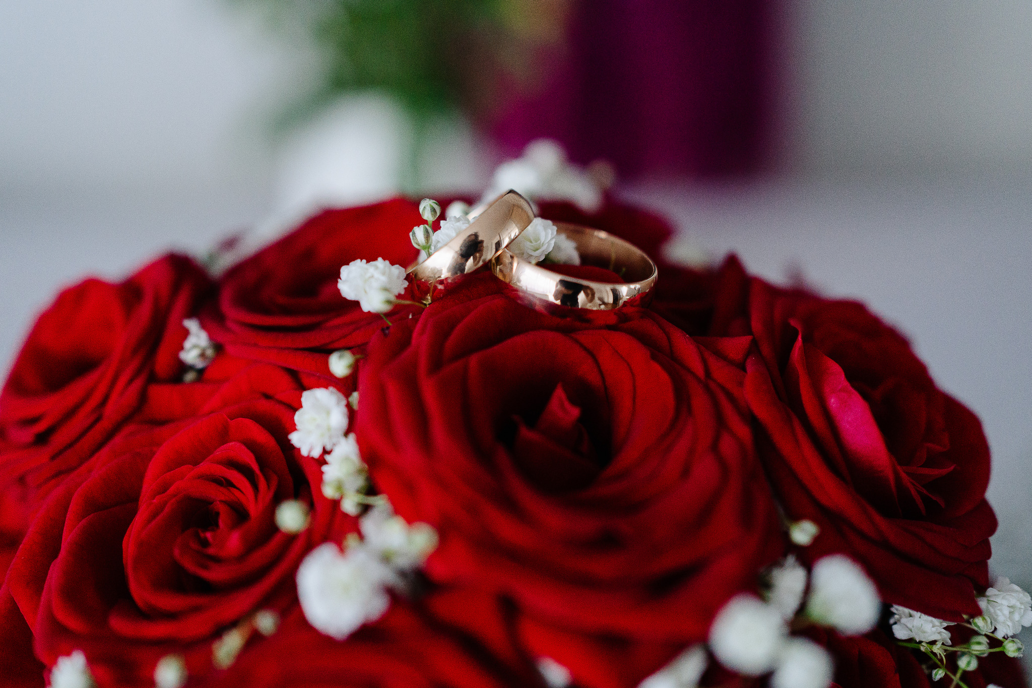 Dwa złote pierścionki ślubne ułożone na bukiecie z czerwonych róż i białych kwiatów gipsówki, w delikatnym, naturalnym oświetleniu.