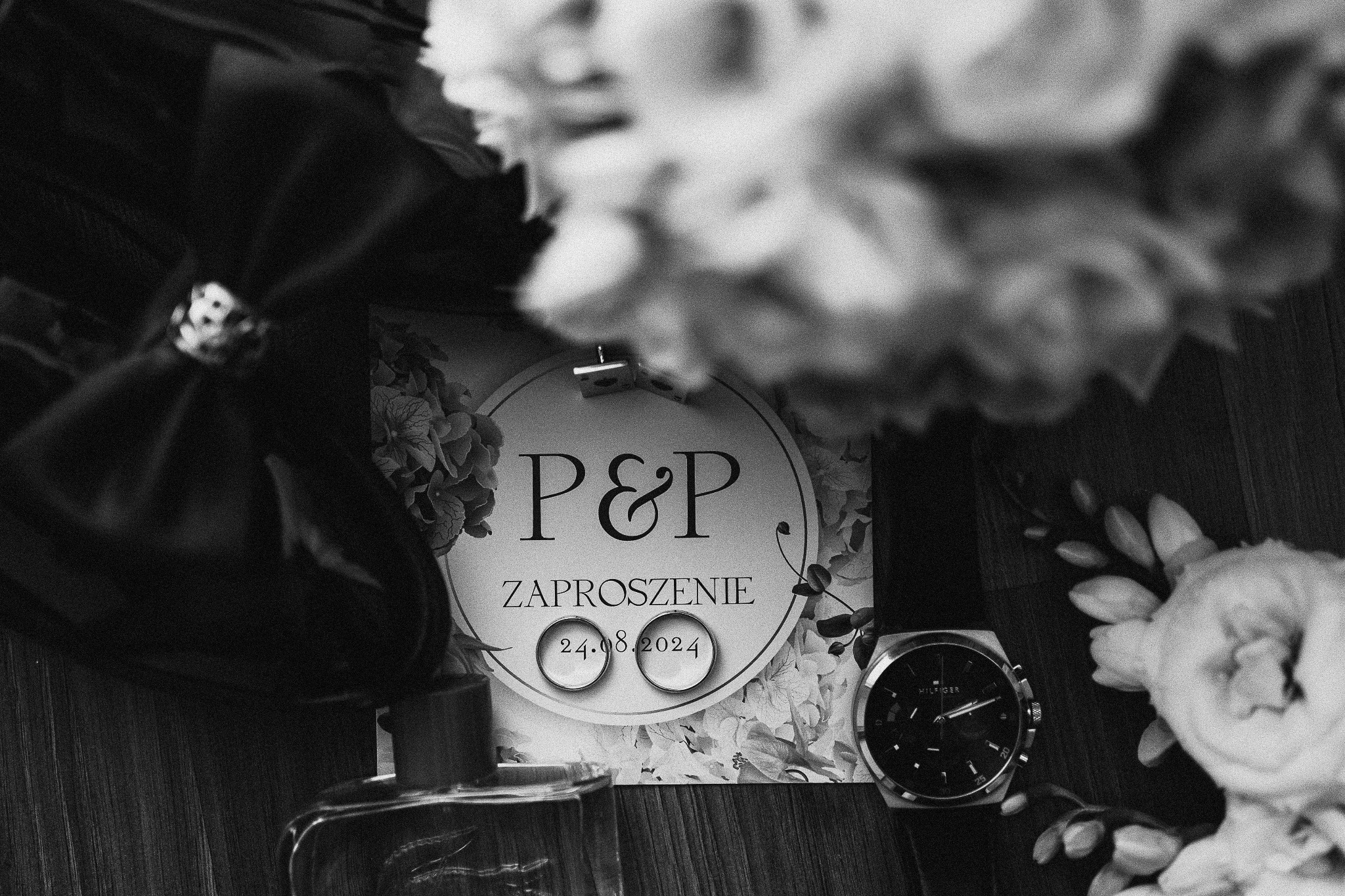 Czarno-białe zdjęcie zaproszenia ślubnego P&P z datą 24.08.2024, obrączek, zegarka Tommy Hilfiger, bukietu i muchy, tworzące kompozycję na drewnianym tle.